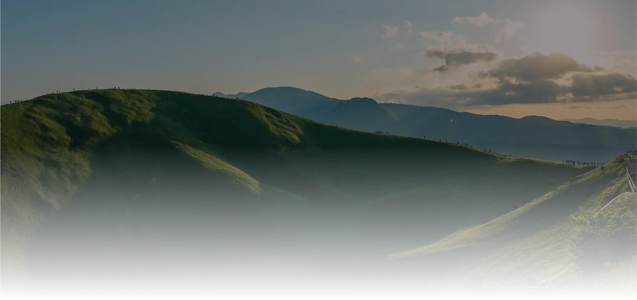 イメージ：夕暮れ時のなだらかな緑の山の稜線に、小さな人々のシルエットが見える風景