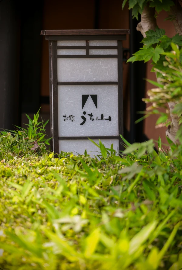 日本の宿の入り口を示す、木と和紙のような素材でできた提灯型の看板。看板には「お宿うち山」という文字と幾何学的な紋様が書かれており、周囲には緑豊かな植物が生い茂っています。