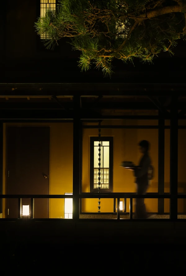 夜、日本の伝統的な宿の廊下を、浴衣を着たスタッフが盆を持って歩いている様子。廊下の奥は明るく照らされ、窓からは障子のような格子が見えます。上部には松の木の枝が写り込み、静かで落ち着いた夜の雰囲気を醸し出しています。