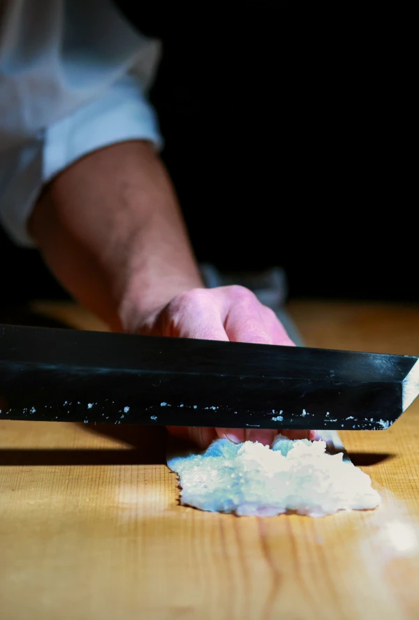 まな板の上で、料理人の手が大きな黒い包丁を使い、白い魚の身を細かく刻んでいる様子を捉えたクローズアップ画像。
