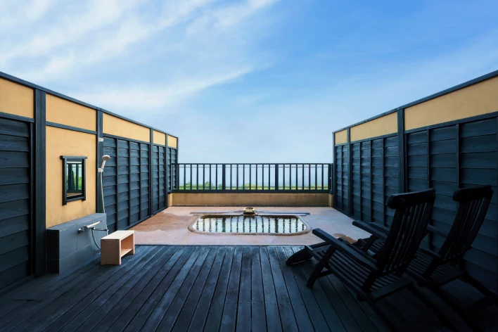 広々としたウッドデッキには、海または広い空を望むことができる場所に石造りの浴槽があり、湯が張られています。浴槽の周囲は低めの塀で囲まれ、左手にはシャワーと鏡が設置されています。右手には黒い木製のリクライニングチェアが2脚置かれており、くつろげる空間が広がっています。