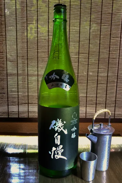 背景の竹簾を背に、深緑色の酒瓶「磯自慢 吟醸」が中央に立っています。瓶の上部には「吟醸 山田錦」と書かれた黒いラベル、中央には「磯自慢」と白く書かれた黒いラベルがあります。手前には銀色の猪口と、取っ手が付いた銀色の小さな徳利が置かれています。