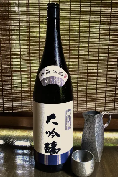 背景の竹簾を背に、黒い酒瓶「磯自慢 大吟醸」が中央に立っています。瓶の上部には「一滴入魂 山田錦」と書かれた白いラベル、中央には「大吟醸」と黒く書かれた白いラベル、下部には青と白のストライプ模様があります。手前には銀色の猪口と、取っ手が付いた銀色の小さな徳利が置かれています。