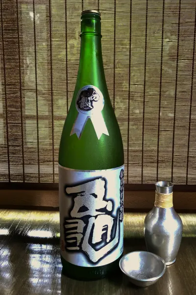 背景の竹簾を背に、深緑色の酒瓶「秘蔵酒 亀 純米大吟醸」が中央に立っています。瓶の上部には白いリボンのような飾りと「亀」の文字、中央には光沢のある銀色で「亀」と大きく書かれたラベルがあります。手前には銀色の猪口と、金色の糸が巻かれた首を持つ銀色の小さな徳利が置かれています。