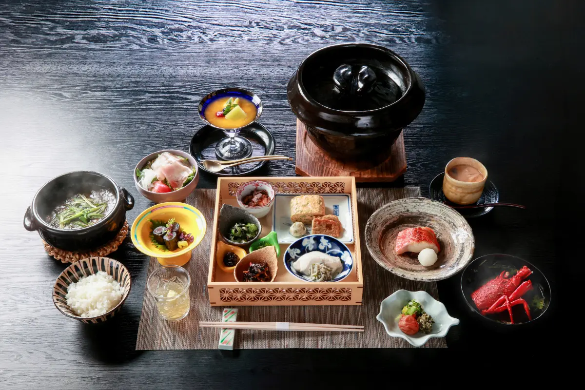 濃い木目のテーブルに、日本の伝統的な朝食の膳が広げられています。中央の木製トレーには、玉子焼き、魚、煮物など様々な小鉢が美しく並べられ、その手前にはご飯が盛られたお椀とグラスに入った飲み物、奥には蓋付きの黒いお鍋、そして湯気の立つ小鍋が見えます。さらに、左奥には黄色いデザート、右側にはカニの身のようなものが盛り付けられた器、そして右手前には可愛らしい形の小鉢が配置され、品数の豊富な食事が全体を覆っています。