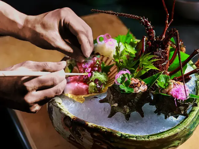  料理人が氷を敷き詰めた器に、伊勢エビの姿造りや数種類の刺身を華やかに盛り付けている様子です。