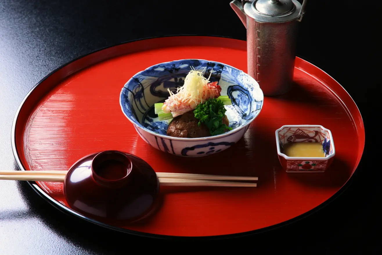 黒い背景に置かれた朱色の丸盆の上に、美しく盛り付けられた冬の料理が並んでいます。中央には、青と白の模様が施された器に、茶色の餡の上にカニの身、緑の野菜、薄切りのネギなどが盛り付けられた温かい料理があります。