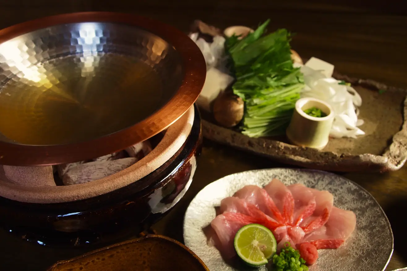 温かい木目のテーブルに、冬の鍋料理の準備が整えられています。手前左には銅色の鍋が置かれ、その右隣には白い皿に美しく盛り付けられた薄切りの魚介と、ライムのような柑橘類、そして紅葉した葉が添えられています。奥には、鮮やかな緑色の葉野菜、きのこ、豆腐、白菜、そして細かく刻まれたネギが入った竹筒など、様々な具材が並べられています。