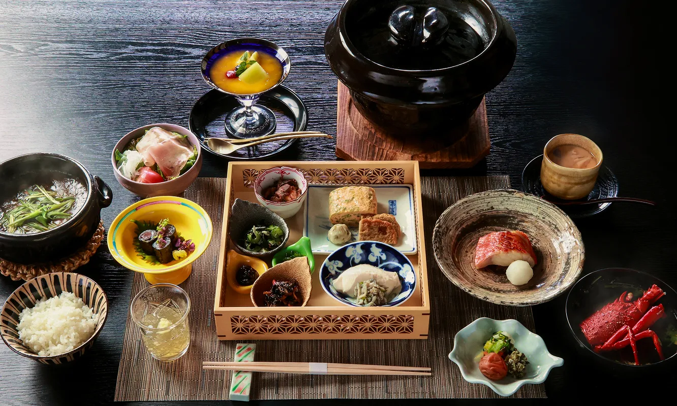 和食の豪華な御膳。中央の木製のお盆には、様々な小鉢に盛られた色とりどりの料理が並び、揚げ物や和え物、煮物などが見える。その周りには、土鍋に入った汁物、ご飯、焼き魚、刺身、サラダ、デザートのフルーツ、飲み物などが美しく配置されている。