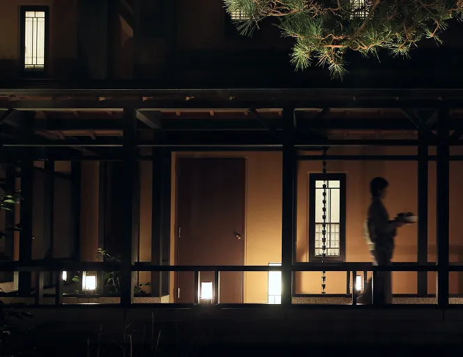 夜の回廊を着物姿のスタッフが料理を運んで歩いている。あたたかな灯りに照らされた和風建築の風情がただよう。